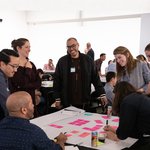 Seven people at a workshop smile around a table as they chat and arrange pink post-its.