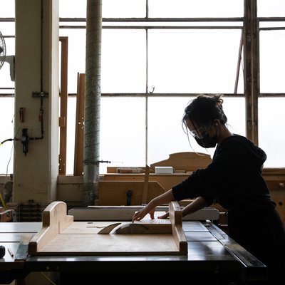 A student cuts wood wearing goggles and a face mask in a light-filled workshop.